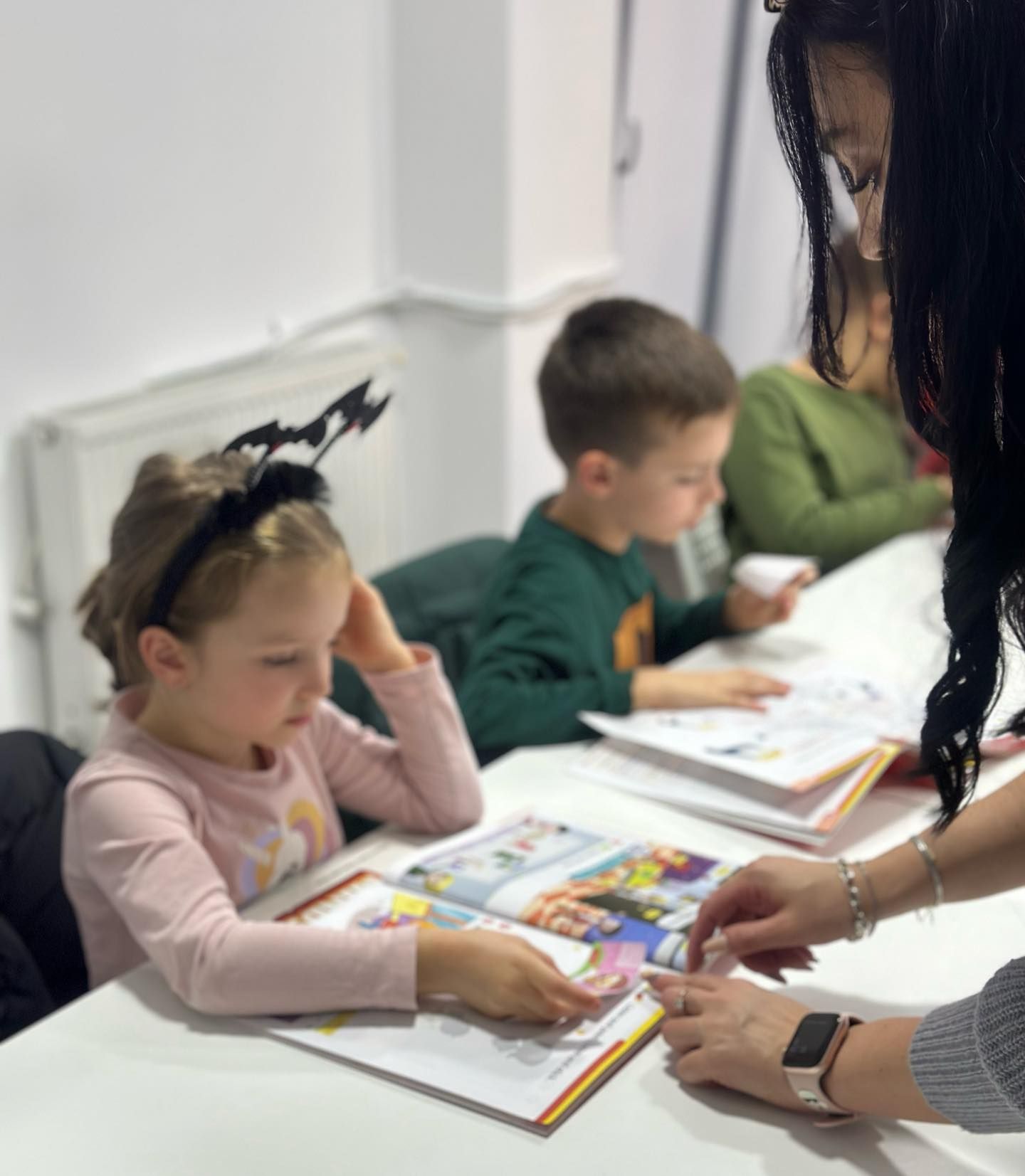 O fetiță și un băiat lucrează în manuale de engleză, iar profesoara se apleacă să le arate ceva.