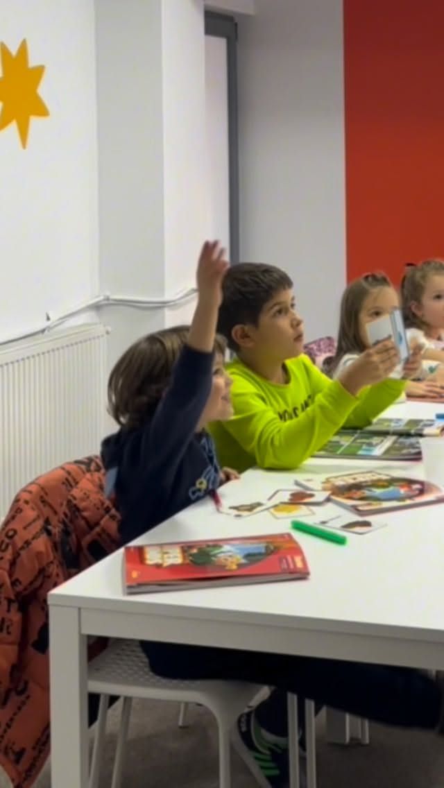 Copii la masă în timpul orei de engleză; unul dintre ei ridică mâna ca să răspundă.
